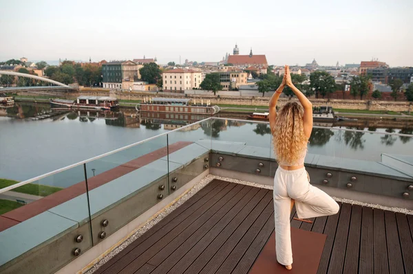 Back view of woman with long curly hair standing at one leg and practicing yoga on wooden terrace with the amazing view to the old Krakow city and river