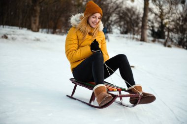 Sarı kışlık ceketli, mutlu, genç bir kadın kar parkındaki kızağın üstünde tepeden aşağı iniyor. Kızak kayan kız