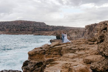 Genç bir düğün çifti, karı koca, deniz kenarındaki kayalıklarda kucaklaşıyorlar. Kıbrıs 'ta romantik bir düğün fotoğrafı çekimi. Yeni evlilerin balayı..