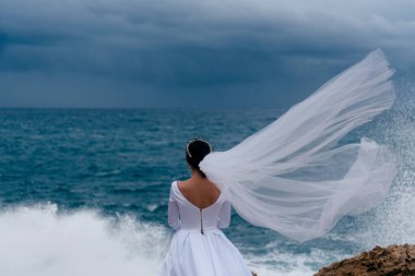 Bulutlu bir günde beyaz gelinlik ve duvak takan esmer gelin. Deniz kenarında dalgalarla poz veren romantik, güzel bir gelin. Gelin duvağı rüzgarda uçuyor.