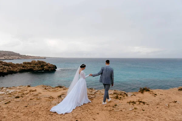 若い結婚式のカップルは青い海の近くの岩場のビーチを歩いています 新郎は花嫁の手を握っています 美しい結婚式の日 — ストック写真