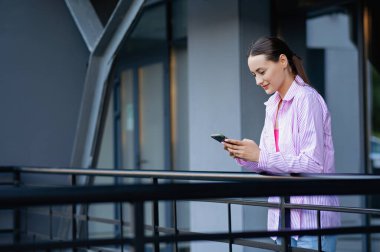 Modern binanın yanında duran ve elinde cep telefonu olan güzel bir kadının yan görüntüsü. Şık kıyafetler içinde internette akıllı telefon sörfü yapan çekici bir esmer. Yaşam Tarzı konsepti.