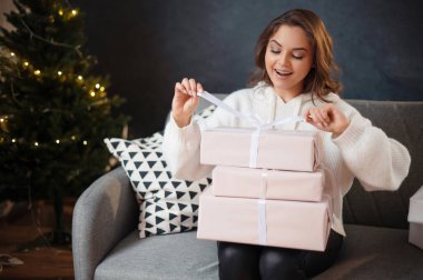 Noel ağacının yanındaki kanepede otururken hediye kutusunda fiyonk bağlayan kıvırcık saçlı neşeli genç bayan. Yeni yıl kavramı