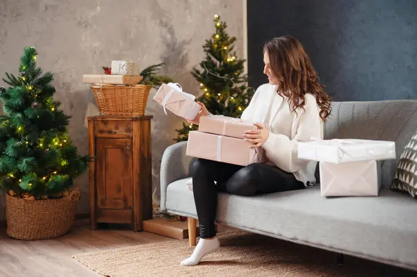 クリスマスツリーの近くのソファに座っている間 贈り物を調べ巻き毛の幸せな若い女性 新年のコンセプト — ストック写真