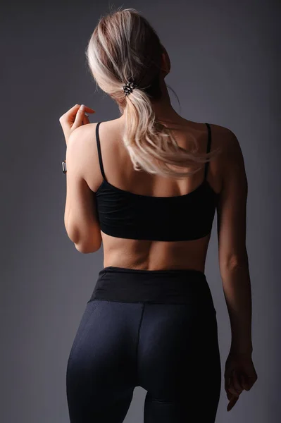 stock image Sporty young woman in sportswear showing her figure on grey studio background. Back view of blonde fitness girl wearing black top and leggings