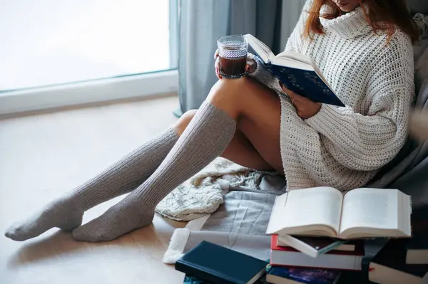 Beyaz örgü süveter ve çorapla yerde kitap ve kahveyle oturan çekici kadın manzarası.