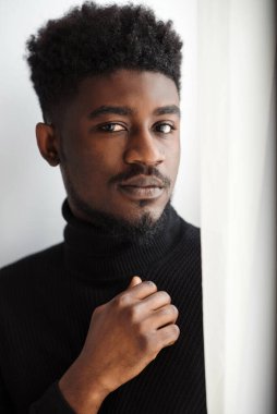 Üzgün siyah adamın kısmi görüntüsü. Depresyondaki genç, kıvırcık sakallı adam kameraya bakıyor. Siyah kazak giy.