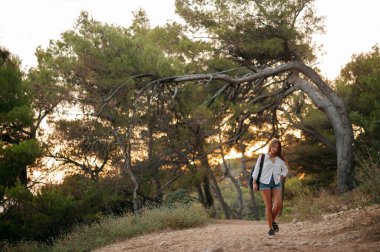Sırt çantalı gezgin kadın gün batımında ormanda yürüyor. Beyaz gömlekli ve kot şortlu bir kız.