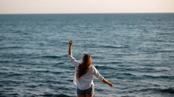 Back view of attractive woman with raised hands in a white shirt and shorts standing near the sea and enjoying the view. Panoramic picture