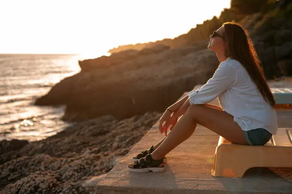 Gün batımında deniz kenarında bir bankta oturan genç bir kadın hayal etmek. Seyahat ve dinlenme konsepti