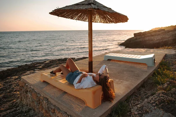 Attractive young woman relaxing on sun lounger near the sea at beautiful sunset. Travel and rest concept