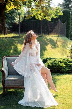 Beyaz sabahlık ve temiz havada antika sandalyeye yaslanmış iç çamaşırlı şehvetli genç bir kadın. Güneşli bir sabahta açık havada poz veren sarı saçlı beyaz bir gelin..