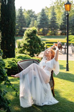 Düğün makyajlı, açık havada sandalyede dinlenen neşeli beyaz bir gelin. Her anın tadını çıkar. Olumlu insan duyguları.