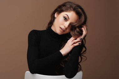 Çekici portresi olan güzel gülümseyen Avrupalı genç bir kadın. Siyah kazak giyen, güzel yüzlü ve saçlı bir kız sandalyeye oturup kameraya bakar. Kahverengi arka planda izole edilmiş. Boşluğu kopyala