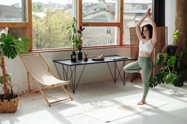 Ağaç yogası yaparken tek ayak üzerinde duran gülümseyen güzel kadın rahat atmosferik bir yerde poz veriyor. Zinde ve narin kadın egzersiz yapıyor ve vrikshasana 'yı beyaz paspasın üzerinde deniyor..