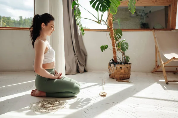Calm and spiritual woman sitting on knees on white yoga mat with closed eyes in bright room. Attractive woman dressed in comfortable sports clothing meditating on floor.