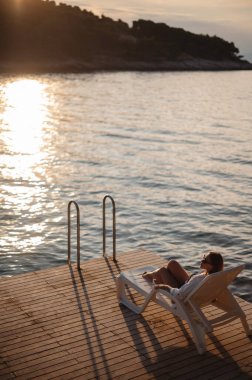 Güneş batarken tahta iskelede, güverte sandalyesinde dinlenen ince tenli kadının yüksek açılı görüntüsü. Güzel esmer, Adriyatik Denizi kıyısında yaz tatilinin tadını çıkarıyor. Eğlence anlayışı..