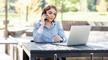Avrupalı iş kadını dışarıda çalışıyor. Güzel genç kız masada oturur, cep telefonuyla konuşur ve dizüstü bilgisayar kullanır. Modern kadın yaşam tarzı. Uzak ve serbest çalışma kavramı