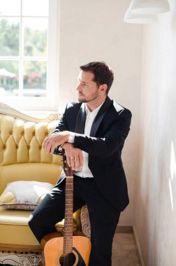 Modern oteldeki kanepede oturan yakışıklı genç adam. Çekici adam gitara yaslanıyor ve başka tarafa bakıyor.