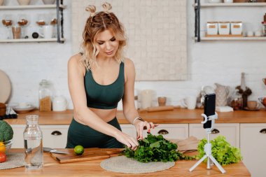 Yeşil spor kıyafetli çekici bir kadın salata kesiyor ve sağlıklı yiyeceklerle ilgili videolar kaydediyor. Mutfakta dikilen sarı saçlı ve özel ders veren fitness blogcusu