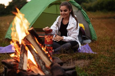 Smiling young woman preparing putting the kettle on the fire. Joyful woman preparing drink outside. Woman drinking a cup of warm tea 