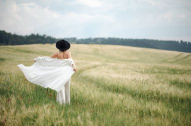 Beyaz elbiseli, siyah şapkalı, tarlada yürüyen güzel sarışın bir kadının arka görüntüsü. Metinleriniz için boşluk kopyala