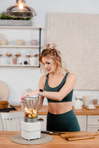 Blender 'da taze meyve suyu içen sportif kadının dikey görüntüsü. Pozitif sarışın kız meyve kokteyli hazırlıyor.