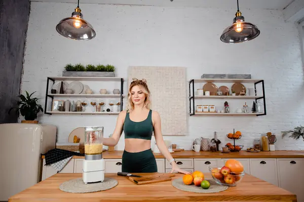 Genç kaslı kadın modern mutfakta dururken taze meyve karıştırıyor. Spor kıyafetli mutlu sarışın idmandan sonra vitamin kokteyli yapıyor..