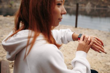 Gölün kenarındaki bankta otururken akıllı saati kullanan genç bir kadın. Sportif kız antrenmandan sonra sonuçlarını kontrol eder.