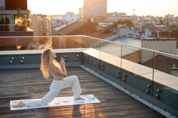Gün batımında çatıda yoga yapan bir kadın. Hamle pozu veriyor. Rahat, açık renkli giysiler giyiyor ve kıvırcık saçları var. Şehrin ufuk çizgisi arka planda görülebilir, sıcak güneş ışığı manzarayı aydınlatır..