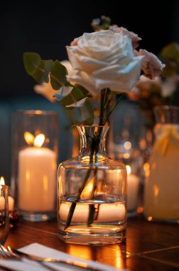 Masadaki cam vazoda beyaz bir gül ve mum ışığında romantik bir ortam..