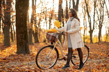 Günbatımında sonbahar parkında bisiklet yürüyüşü yapan mutlu aktif genç kadın. Sonbahar yapraklarıyla dolu sepeti olan pozitif bir kız. 