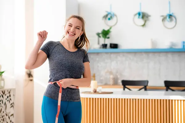 Spor yapan mutlu beyaz kadın, mutfak adasının yakınında dururken bel ölçme bandıyla elini havaya kaldırıyor. Evde başarının zayıflatılması konusunda heyecanlanan duygusal bir kadın..