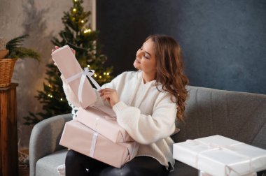 Noel ağacının yanındaki kanepede otururken hediye kutusunda fiyonk bağlayan kıvırcık saçlı neşeli genç bayan. Yeni yıl kavramı