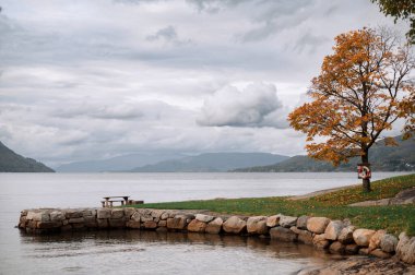 Taş duvarlı sakin bir göl manzarası, sonbahar yapraklı bir ağaç ve piknik masası. Bulutlu gökyüzü ve uzak dağlar sakin bir atmosfer yaratır..