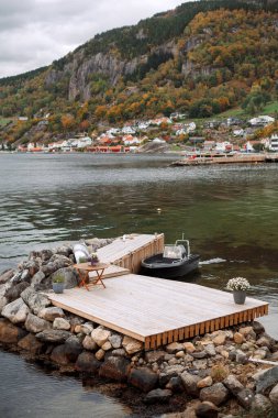 Huzurlu bir göl manzarası, ahşap bir rıhtım, küçük bir tekne ve kayalık bir sahil. Arka planda, renkli bir sonbahar yeşilliği ve bulutlu bir gökyüzünün altında tuhaf bir köy görülebilir..