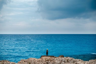 Kayalık sahilde tek başına duran, bulutlu bir gökyüzünün altındaki engin okyanusa bakan bir figür. Sahne yalnızlık ve tefekkür hissini yakalar..