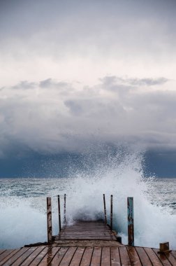 Dalgaların bulutlu bir gökyüzünün altında çarptığı çalkantılı bir denize doğru uzanan ahşap bir iskele..