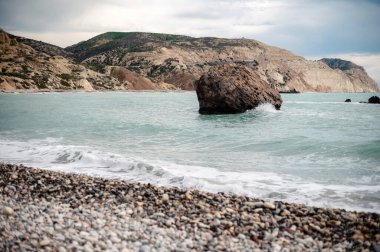 Turkuvaz suları ve bulutlu bir gökyüzünün altındaki uzak tepelerle çevrili, denizde büyük bir kaya parçasıyla kayalık bir sahil manzarası..
