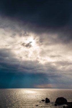 Kara bulutlar ve güneş ışınlarıyla okyanus yüzeyini aydınlatan dramatik deniz manzarası. Kayalar ön planda görünür, sakin ama dengesiz bir atmosfer yaratır..