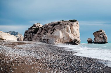 Bulutlu bir gökyüzünün altında büyük kaya oluşumları ve çakıl taşlarıyla manzaralı bir sahil. Dalgalar yavaşça kıyıya çarpar ve sakin bir atmosfer yaratır..