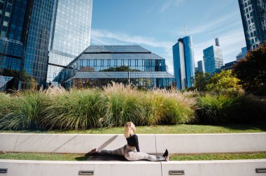 Fitness kadını, arka planda gökdelenler olan bir şehir parkındaki beton bir çıkıntıda ikiye bölünmüş bir performans sergiliyor..