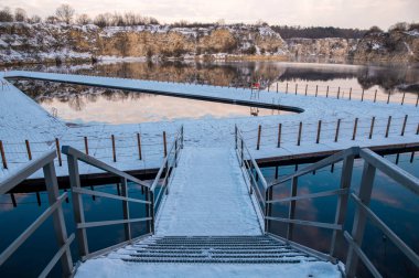 Snow-covered walkway leading to a frozen lake surrounded by trees. clipart