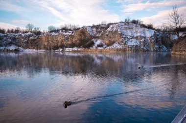 Ördeklerin yüzdüğü sakin bir göl içeren sakin bir kış manzarası. Karla kaplı uçurumlar ve çıplak ağaçlar suyun etrafını sarar. Yumuşak mavi gökyüzünü ve bulutları yansıtır..