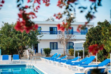 Akdeniz tarzı bir villa, mavi aksanlı, yemyeşil bir yer ve güneş şezlonglarıyla çevrili, canlı kırmızı çiçeklerle çerçevelenmiş bir yüzme havuzu..