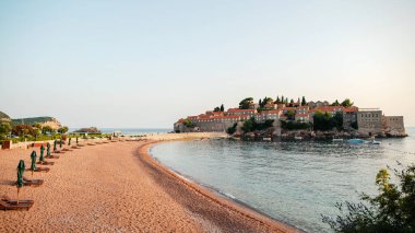 Güneşli şezlongları ve şemsiyeleri olan kumlu bir sahil manzarası, sakin deniz sularıyla çevrili, kırmızı kiremitli ve taştan yapılmış tarihi bir ada köyüne götürüyor..