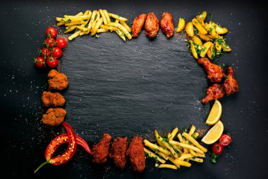 Kızarmış tavuk kanatları, patates kızartması ve sebzeler dahil olmak üzere çeşitli yiyeceklerle çevrili siyah bir tabakta domates ve garnitür olarak limon dilimleri var..