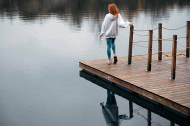 Spor elbiseli kızıl saçlı kadın elinde su şişesi olan sakin bir gölün yanındaki ahşap rıhtımda duruyor. Sahne sakin ve yansıtıcı, su bulutlu gökyüzünü yansıtıyor..