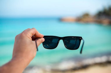 Arka planda tropikal plajı ve turkuaz denizi olan güneş gözlüğü tutan bir el..