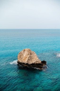 Bulutlu bir gökyüzünün altında berrak mavi okyanus suyuyla çevrili ıssız bir kaya oluşumu..